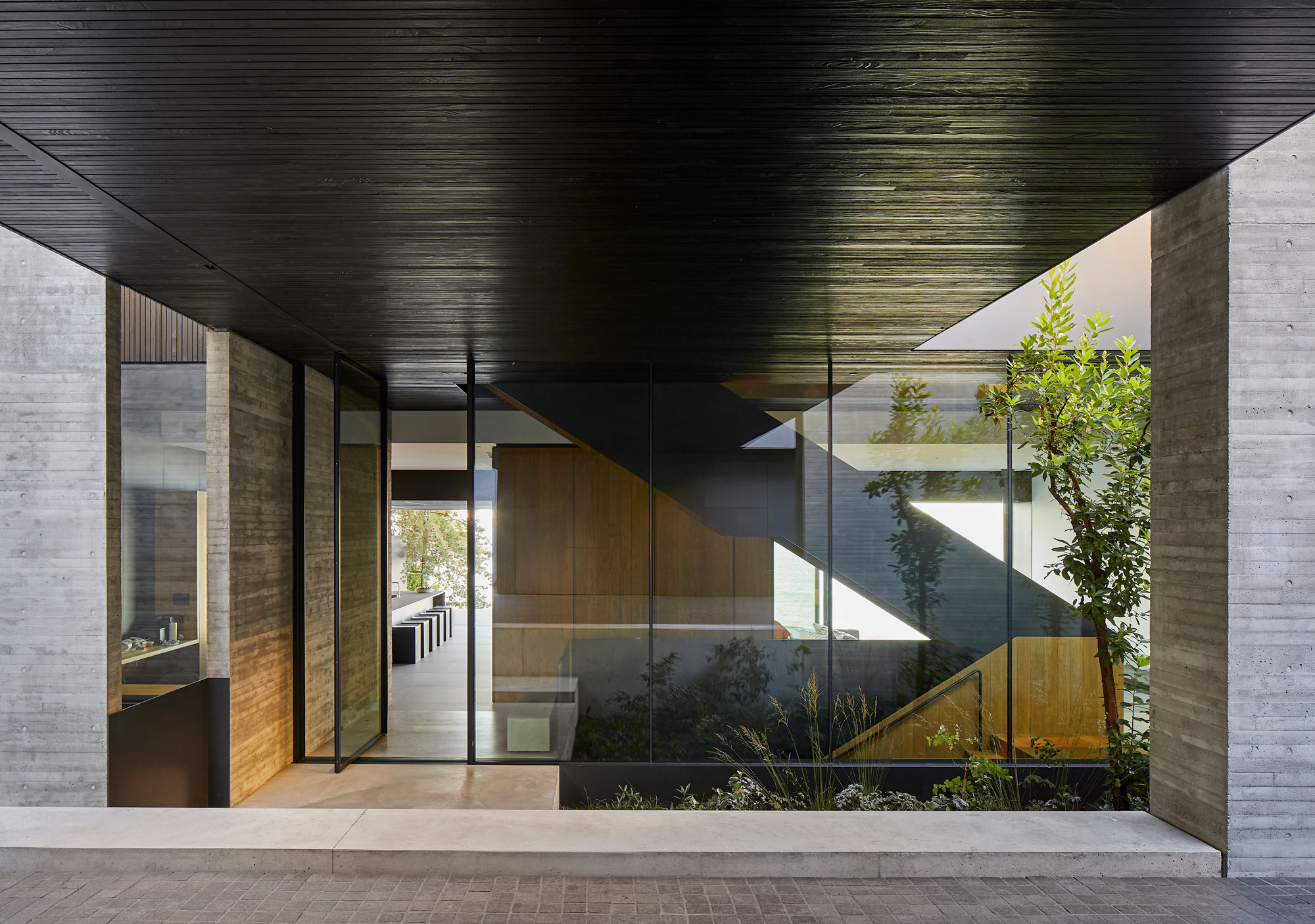 Modern building entryway with large glass walls, concrete surfaces, and a view of an interior stairway and landscaped courtyard.