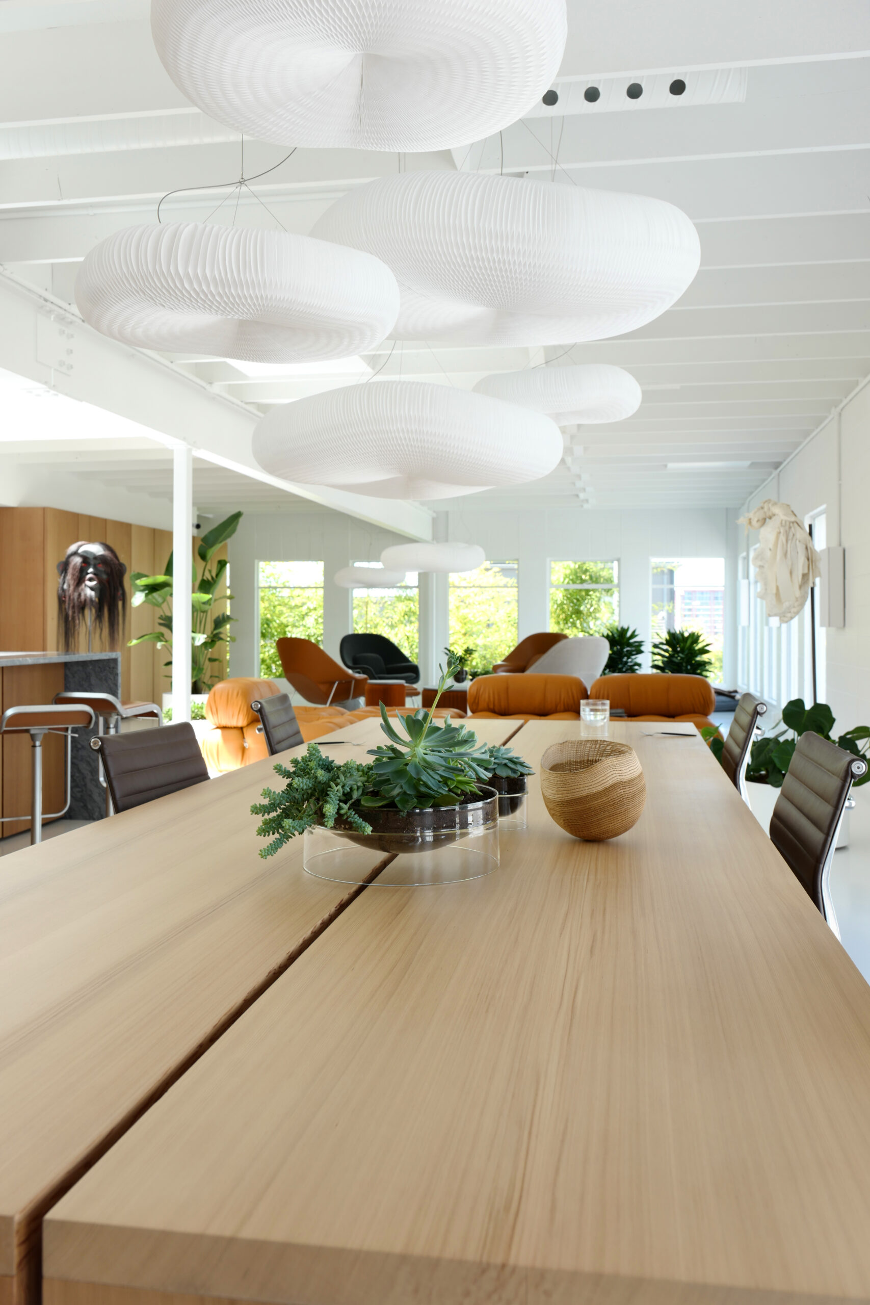 Modern meeting room with a long wooden table, brown chairs, potted plants, decorative vases, and large white pendant lights. Orange armchairs and greenery in the background.
