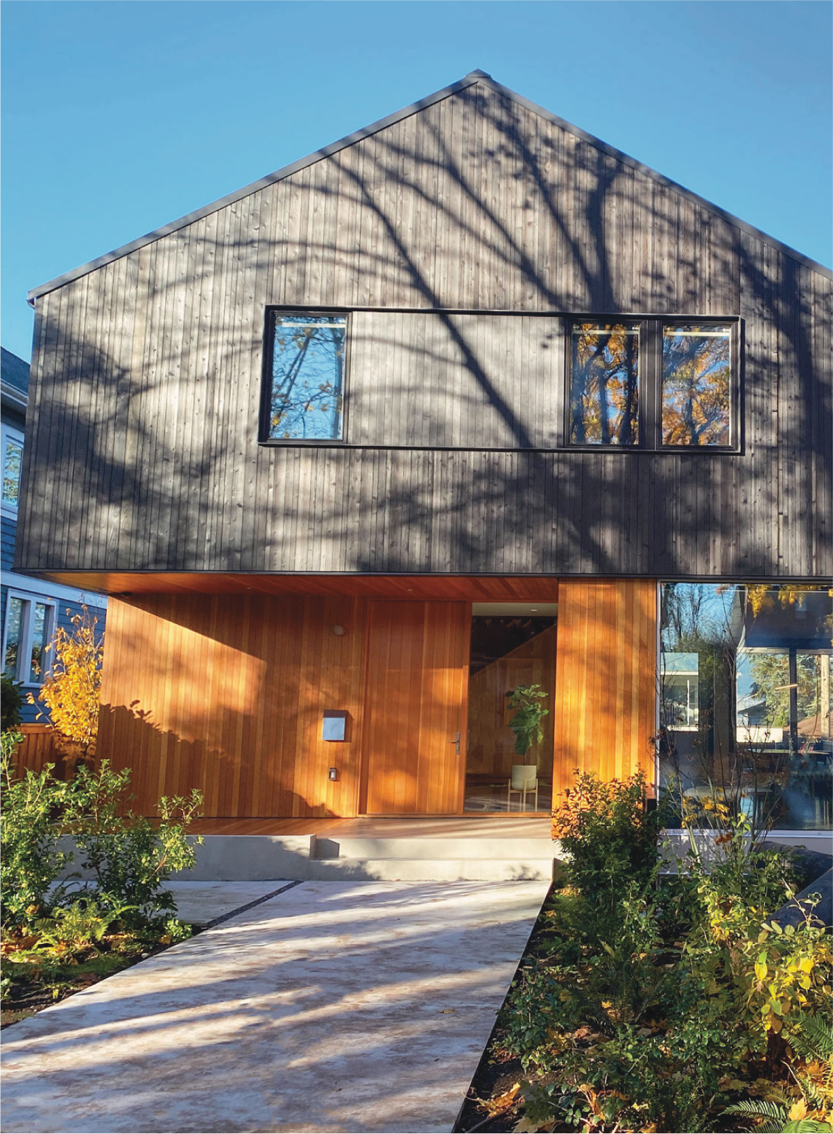 A modern two-story house with a wooden exterior, large windows, and a concrete walkway leading to the entrance, surrounded by landscaping and trees casting shadows on the facade.