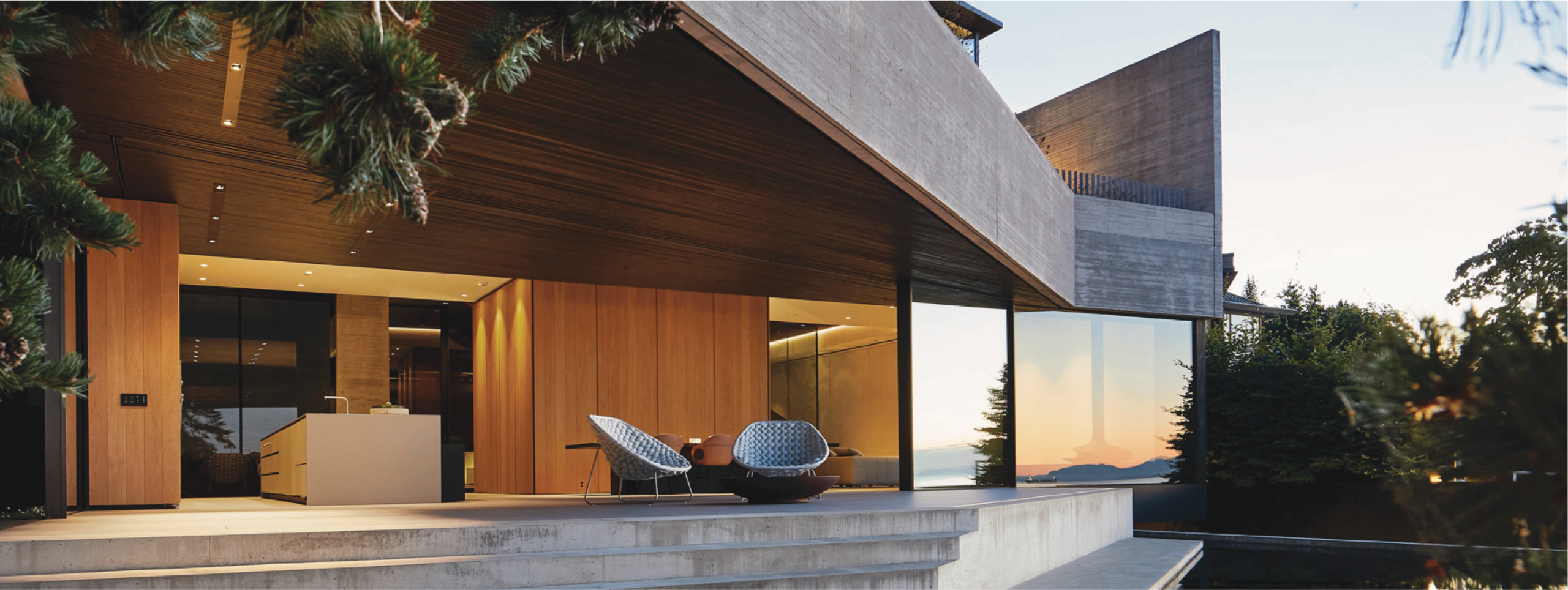 Modern house with large glass walls, wood and concrete exterior, angular roof, and outdoor seating, overlooking trees and a sunset.