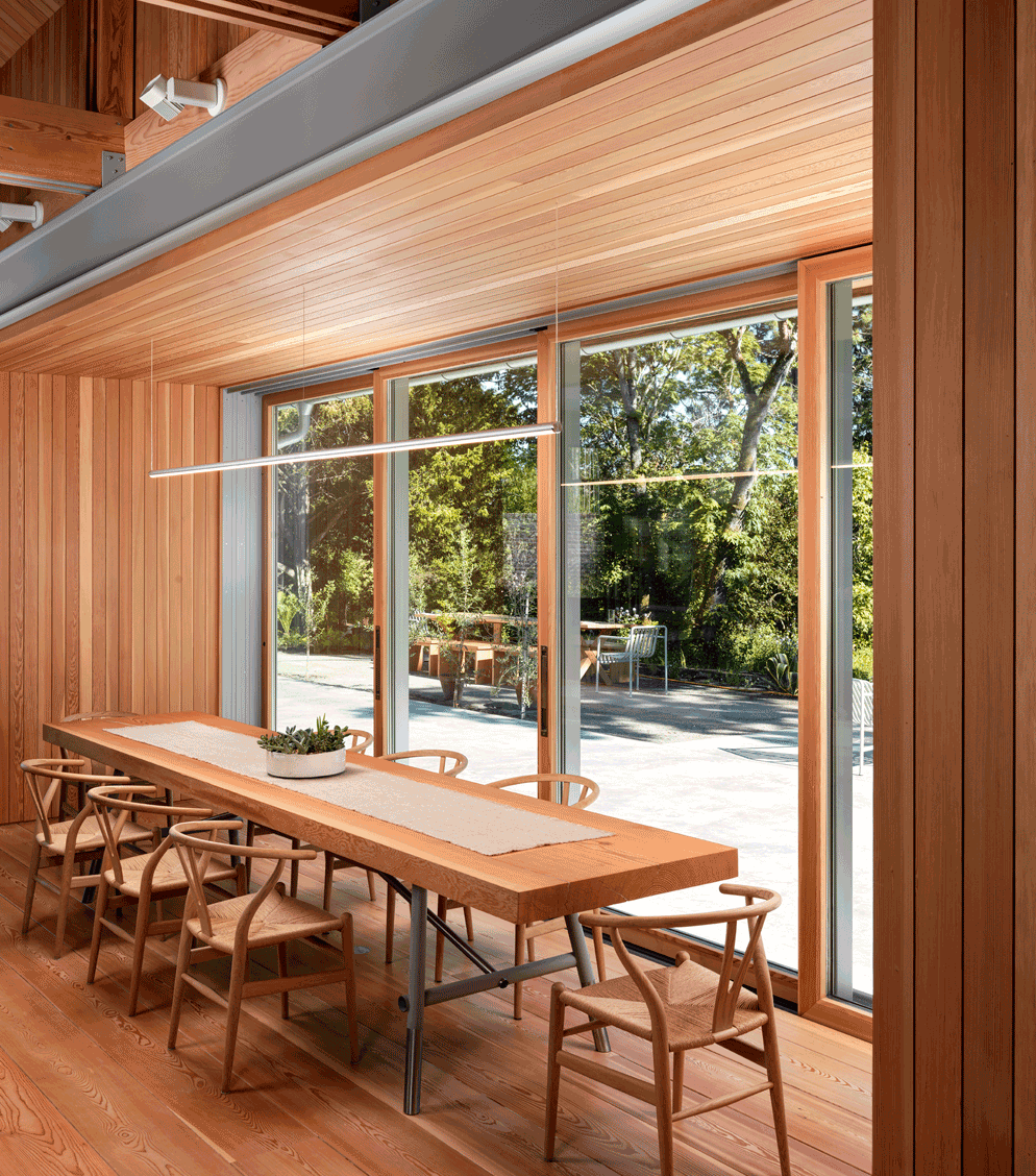 A modern dining area with a long wooden table, matching chairs, and large windows overlooking a patio and greenery outside.