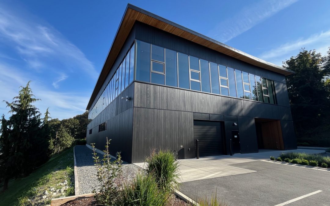 Modern two-story building with large windows, dark exterior panels, and a sloped roof, set against a clear blue sky with landscaped greenery and a paved parking area.
