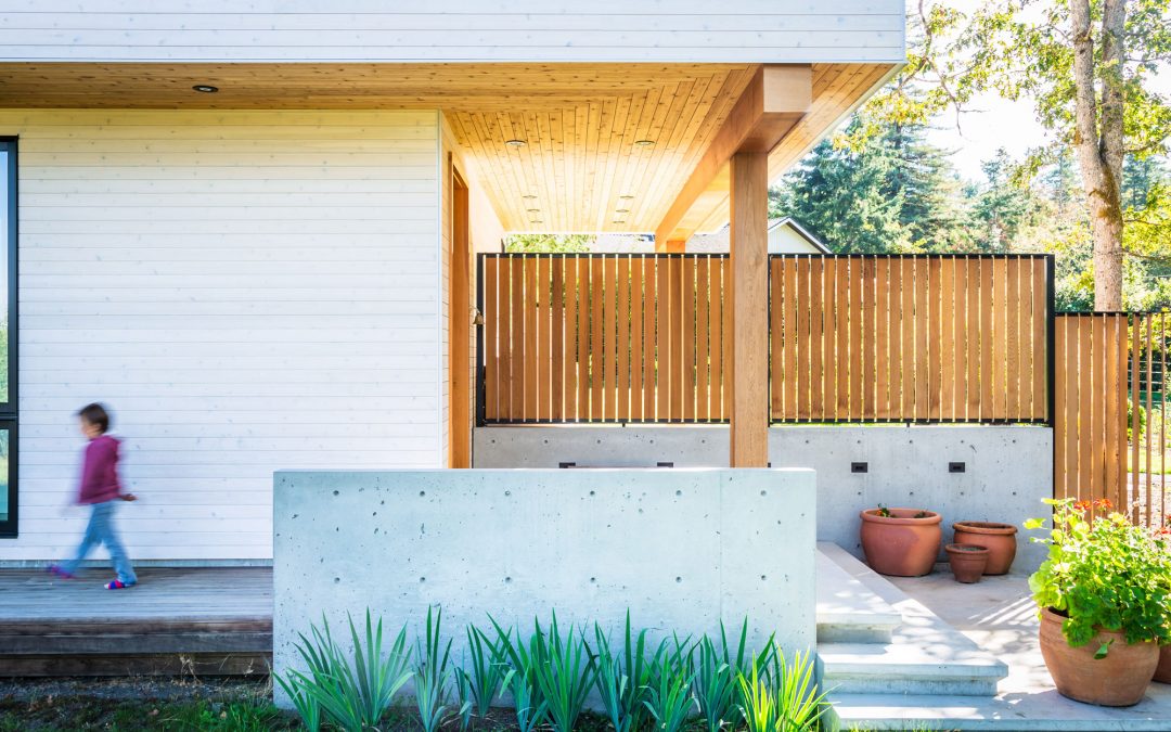 A child walks past a modern house with white wooden walls, a concrete planter, potted plants, and a wooden privacy fence.