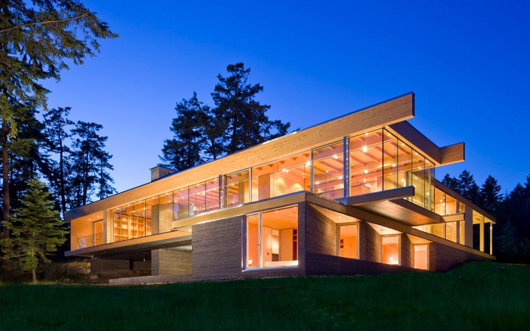 Modern two-story house with large glass windows and wooden beams, illuminated at dusk, surrounded by tall trees and greenery.