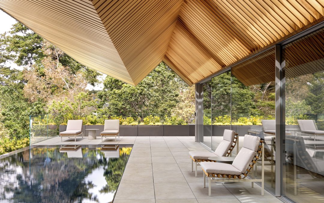 Modern outdoor pool area with cushioned lounge chairs, a reflecting pool, large glass windows, and an overhanging wooden slatted roof, surrounded by greenery.