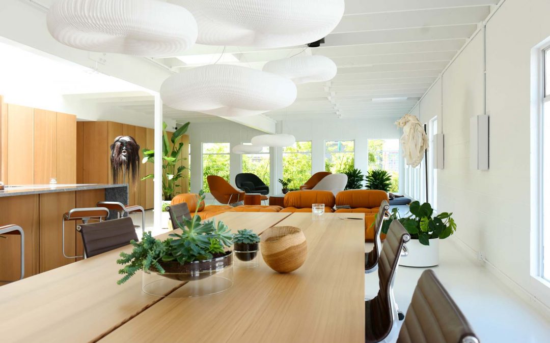 A modern, bright office space features a large wooden table with plants, contemporary chairs, lounge seating, oversized white pendant lights, and large windows letting in natural light.