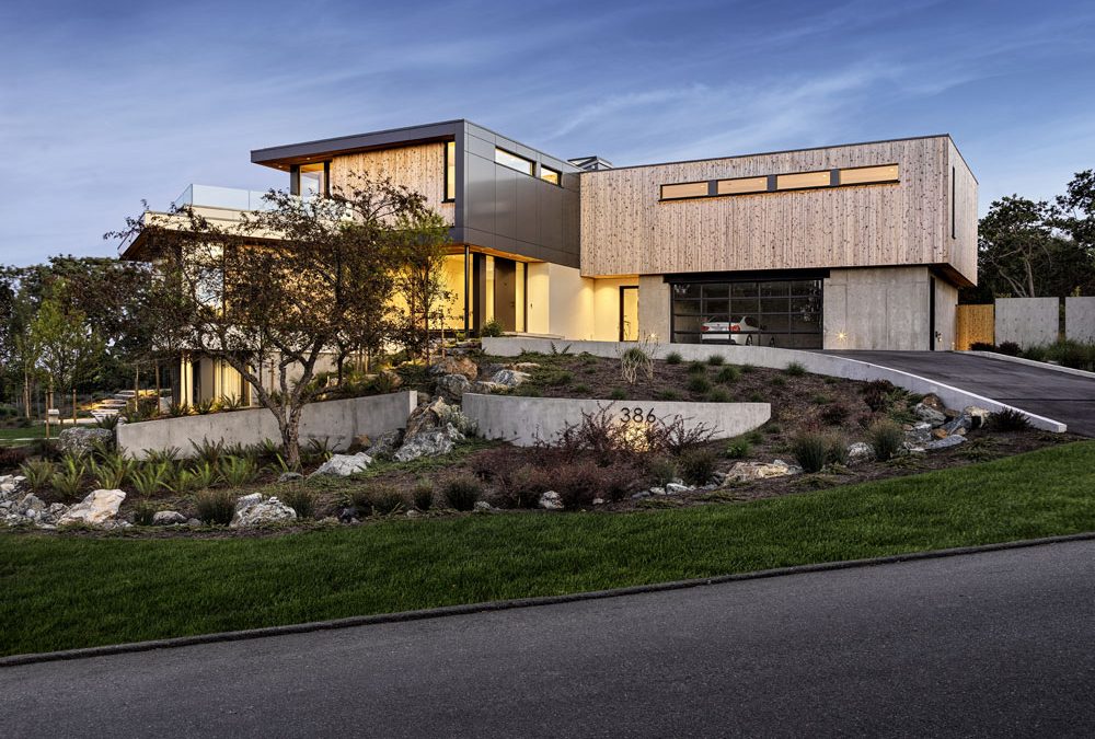 Modern two-story house with large windows, wood and concrete exterior, landscaped yard, driveway, and visible house number 386 at dusk.