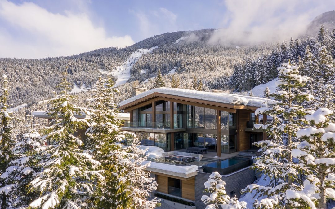 Modern house with large windows and terraces surrounded by snow-covered pine trees, set at the base of a mountain under a partly cloudy sky.