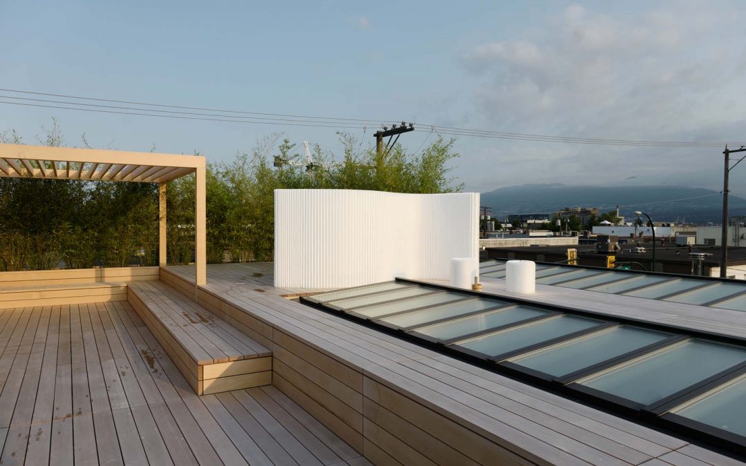Rooftop deck with wooden flooring, built-in seating, pergola, glass skylights, and partial white privacy screen; urban and mountain views in the background.