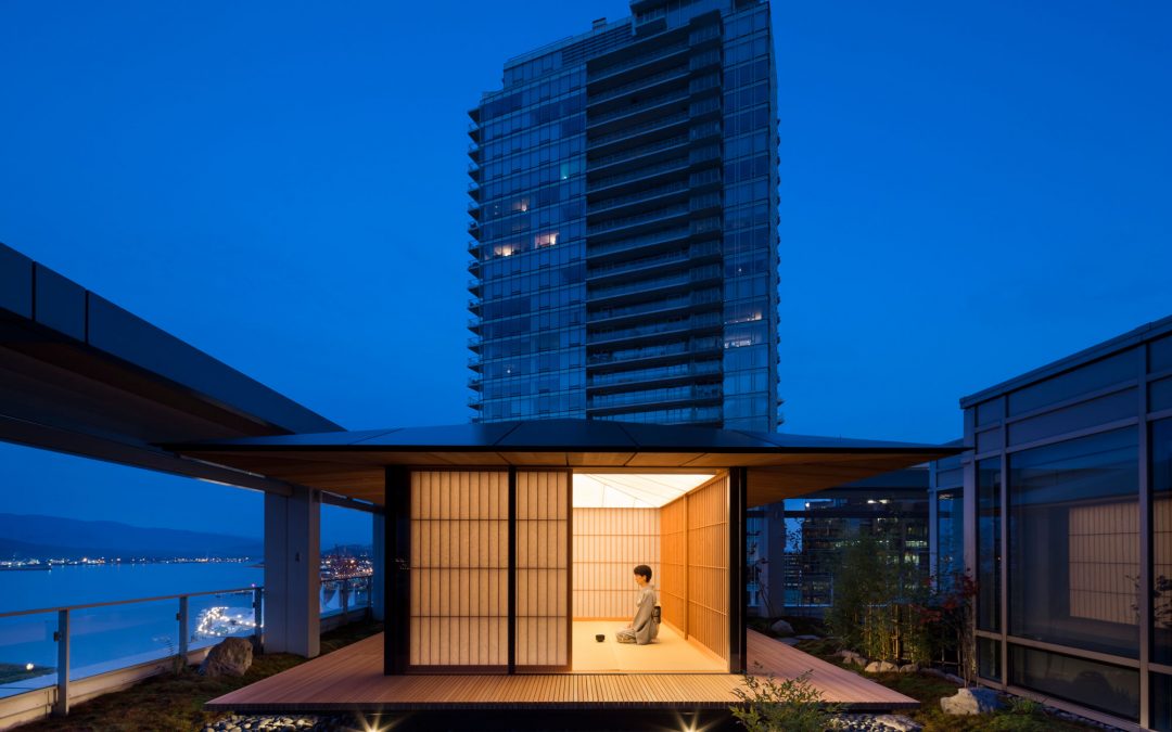 A person sits inside a modern, illuminated tea house on a rooftop at dusk, with a tall glass building and cityscape in the background.