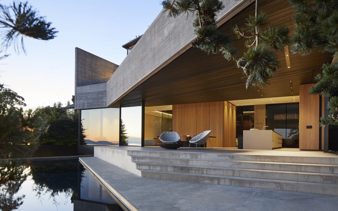 Modern house with large glass windows, wood accents, and concrete structure, set beside a reflective pool and surrounded by trees at sunset.
