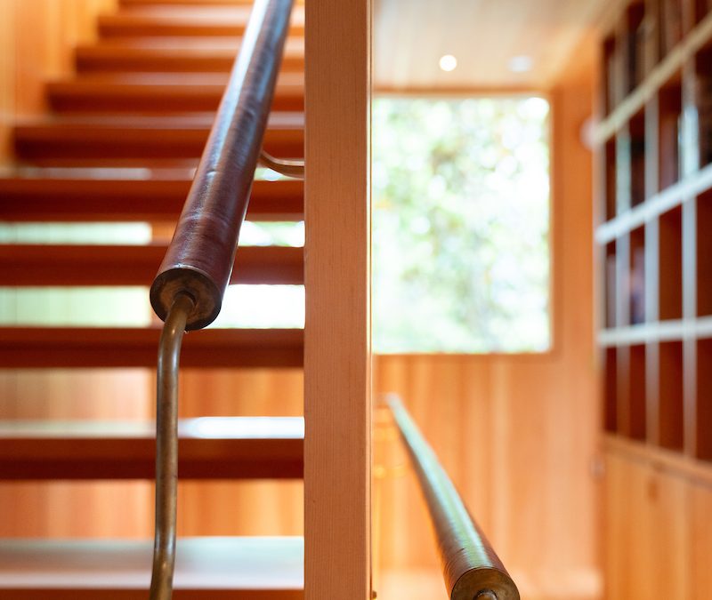 A modern wooden staircase with a metal handrail, illuminated by natural light from a large window, and shelves lining the hallway.