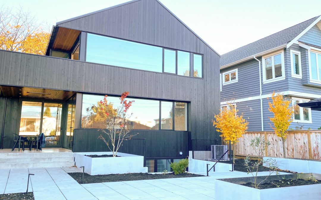 Modern two-story house with dark wood exterior, large windows, and minimal landscaping in the front yard; neighboring house visible on the right.