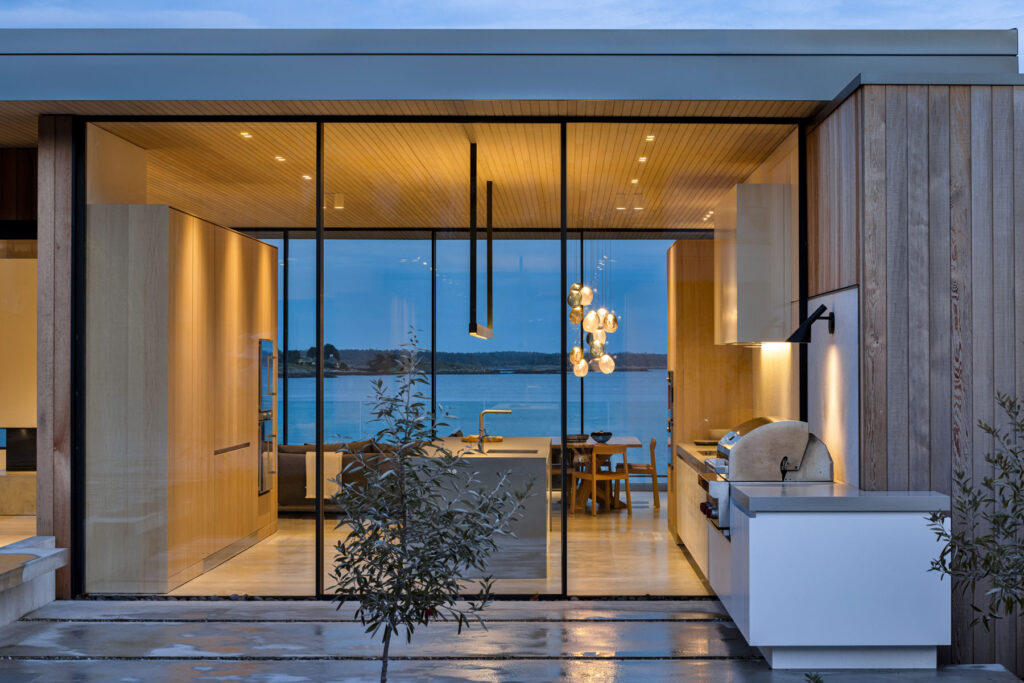 Modern house with large glass doors, wooden exterior, and outdoor kitchen area, overlooking a body of water at dusk. Indoor lighting highlights the open kitchen and dining space.